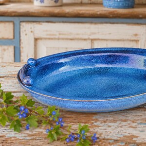 Könnte beinhalten: Eine ovale Servierschale aus Keramik mit einer leuchtenden, glänzenden kobaltblauen Glasur. Die Schale hat ein glattes, abgerundetes Inneres und ein markantes Griffdesign auf der linken Seite, bestehend aus zwei kleinen, kugelförmigen Knöpfen.