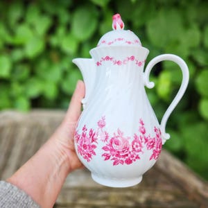 Könnte beinhalten: Weiße Porzellan-Kaffeekanne mit Deckel, verziert mit einem rosa Blumenmuster. Die Kanne hat einen gebogenen Griff und einen Ausguss. Der Deckel ist mit einer rosa Knospe gekrönt. Das Blumenmuster umfasst Rosen und Blätter.