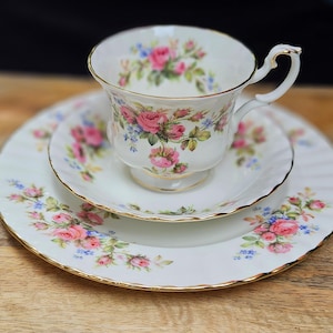 Könnte beinhalten: Ein weißes Porzellan-Teeservice mit einer Tasse, Untertasse und einem Teller. Das Set ist mit rosa Rosen, blauen Blumen und grünen Blättern verziert. Die Ränder sind goldfarben verziert. Die Tasse hat einen gebogenen Henkel.