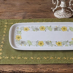 Könnte beinhalten: Ein rechteckiger weißer Servierteller mit einem Blumendesign in Gelb, Grün und Grau. Die Blumen und Blätter umranden die Ränder des Tellers. Der Teller liegt auf einem grünen Läufer mit gestickten gelben Blumen.