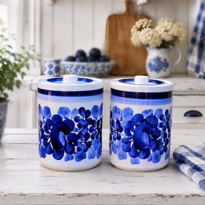 Könnte beinhalten: Zwei zylindrische Keramikdosen mit weißem Grund und handgemalten blauen Blumenmustern. Jede Dose hat einen passenden Deckel mit einem kleinen Knauf, ein breites blaues Band am oberen Rand und ein wiederkehrendes dunkelblaues Blütenmuster.
