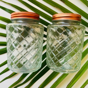 May include: Two clear glass jars with copper lids and a diamond pattern. The jars are empty and sitting on a green leafy background.