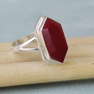 Pode incluir: Um anel de prata com uma grande pedra preciosa vermelha escura, facetada e retangular. A pedra preciosa é engastada em uma moldura de prata, e o anel tem um design de haste dividida. O anel é exibido em uma superfície de madeira marrom claro.