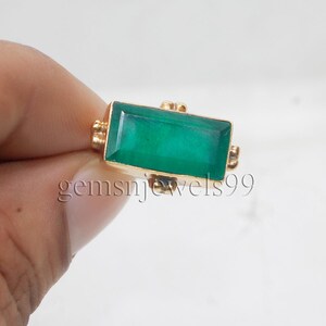 May include: A gold ring featuring a rectangular, faceted green gemstone set horizontally. The metal band has small decorative accents on the sides of the setting. The text gemsnjewels99 is visible across the center of the ring's gemstone.