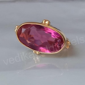 Könnte beinhalten: Ein Ring mit einem ovalen rosafarbenen Edelstein in einer goldenen Fassung. Der Ring hat ein schlichtes Design mit einer glatten, polierten Oberfläche.