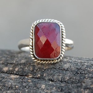 May include: A silver ring with a red gemstone set in a square bezel. The bezel has a rope-like design around the edge.