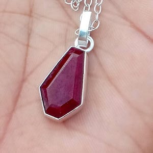 May include: A silver pendant necklace with a faceted, deep red gemstone. The gemstone is set in a silver bezel and hangs from a delicate silver chain. The pendant has a unique, geometric shape.