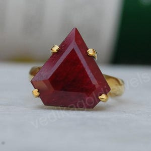 May include: A gold ring featuring a large, faceted, deep red gemstone. The gemstone is cut into a geometric shape with a pointed top. The ring's band and the prongs holding the stone are gold.