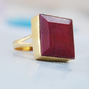 May include: A gold-toned ring featuring a large, square, deep red gemstone. The gemstone is set in a gold bezel, and the band has a simple, elegant design. The ring is displayed on a light-colored surface.