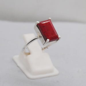Peut inclure: Une bague en argent ornée d'une pierre rouge foncé de forme rectangulaire. La pierre est sertie dans un anneau en argent, créant un contraste saisissant. La bague est présentée sur un support blanc.