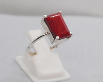 Peut inclure: Une bague en argent ornée d'une pierre rouge foncé de forme rectangulaire. La pierre est sertie dans un anneau en argent, créant un contraste saisissant. La bague est présentée sur un support blanc.