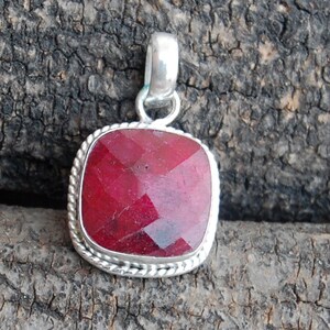 Puede incluir: Un colgante cuadrado con una piedra preciosa roja y un engaste de plata. El colgante tiene un borde de cuerda alrededor de la piedra preciosa.