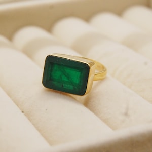 Könnte beinhalten: Ein goldener Ring mit einem rechteckigen, smaragdgeschliffenen grünen Edelstein. Der Ring hat ein schlichtes, elegantes Design mit einem polierten Goldband. Der Edelstein ist in einer goldenen Lünette gefasst.