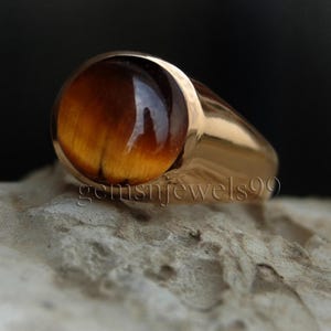 May include: A gold ring featuring a large, round, brown tiger's eye gemstone. The ring has a plain, polished band.