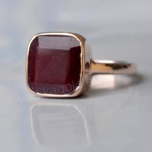 Può includere: Un anello con rubino di forma quadrata in oro rosa. L'anello ha una fascia semplice e un grande rubino sfaccettato.