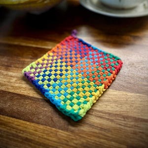 Puede incluir: Un salvamanteles cuadrado tejido en colores del arcoíris, incluyendo rojo, naranja, amarillo, verde, azul y morado. El salvamanteles está hecho de un material grueso y texturizado y tiene un lazo para colgar. El fondo es una superficie de madera.