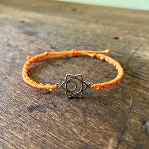 May include: Orange string bracelet with a silver charm in the shape of a chakra symbol.