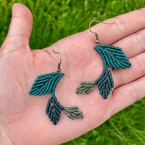 May include: A pair of green and blue macrame earrings with a leaf design. The earrings are made with a dark green and light green cord and have a bronze hook.