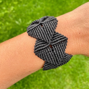 May include: A black macrame bracelet is shown on a person's wrist. The bracelet is made of woven black cord, featuring a geometric design. The background is a blurred green lawn.