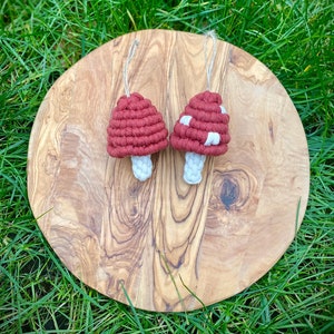 May include: Two crocheted mushroom ornaments with white and red yarn. The mushrooms are hanging from a wooden board.