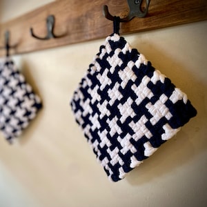 Może przedstawiać: Dwa tkane podkładki pod garnki z czarno-białym wzorem w pepitkę wiszą na drewnianym wieszaku z metalowymi haczykami. Podkładki pod garnki są kwadratowe i mają pętlę do zawieszania. Tło to jasnobeżowa ściana.