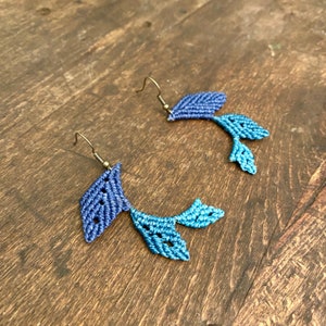 Puede incluir: Un par de pendientes de macramé hechos a mano. Cada pendiente presenta un diseño en forma de hoja en tonos azules, con una parte superior azul más oscura y una parte inferior azul más clara. Los pendientes tienen ganchos para las orejas de color bronce.