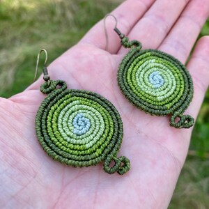Op de afbeelding: Een paar groene en witte gehaakte oorbellen met een spiraalvormig ontwerp. De oorbellen zijn gemaakt met verschillende tinten groen en wit garen, waardoor een verloop ontstaat.
