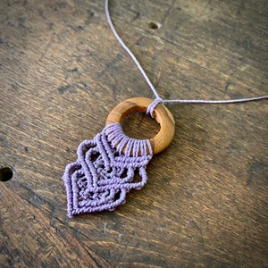 May include: A handcrafted macramé necklace with a wooden ring pendant and detailed lavender knotting. The necklace is presented on a rustic wooden surface, highlighting the natural wood grain and the delicate craftsmanship of the jewellery.