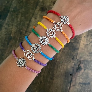 May include: Seven chakra bracelets in a rainbow of colours: red, orange, yellow, green, blue, purple, and pink. Each bracelet has a silver-coloured charm with a unique geometric design. The bracelets are made of woven cord.