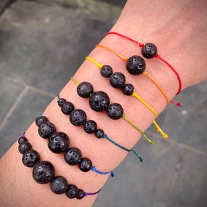 May include: Six bracelets featuring black lava beads on colourful cords. Each bracelet has a different cord colour: red, orange, yellow, green, blue, and purple. The lava beads have a porous texture. The bracelets are displayed in a row.