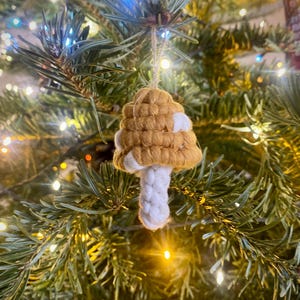 May include: A crocheted mushroom ornament in shades of brown and white hangs on a Christmas tree with white fairy lights.