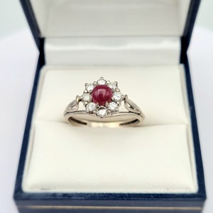 May include: A white gold ring with a red ruby gemstone surrounded by a halo of smaller diamonds.