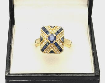 Può includere: Un anello in oro con una testa rettangolare caratterizzata da uno zaffiro blu ovale centrale, circondato da un motivo geometrico di zaffiri blu taglio baguette incastonati a binario e piccoli diamanti bianchi rotondi su una fascia in oro.