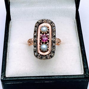 Op de afbeelding: Antieke ring met een rechthoekig ontwerp, met twee parels en een robijn in een centrale rij. De ring heeft een gouden band en een zilverkleurig frame met kleine accentstenen. Gepresenteerd in een zwarte sieradendoos.