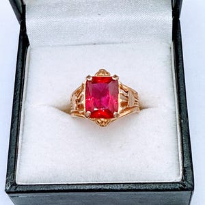 Puede incluir: Un anillo de oro con una gran piedra roja cuadrada engastada en un diseño de filigrana.