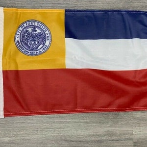 May include: A flag with a yellow, red, white, and blue design. The flag has a seal in the upper left corner that says "City of Fort Smith Ark. Incorporated A.D. 1842".