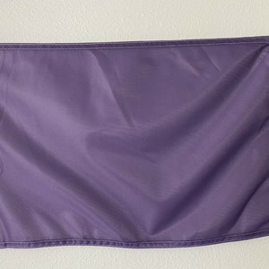 May include: A solid purple flag with white stitching around the edges. The flag is hanging from a white wall with a silver clip.