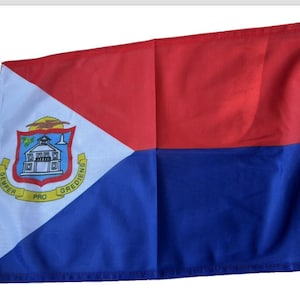 May include: A blue, white, and red flag with the coat of arms of Sint Maarten in the upper left corner. The coat of arms features a building with a tower, a bird, and the words "SEMPER PRO GREDIENS".