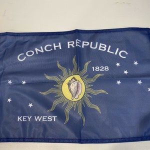 May include: A blue flag with the words "Conch Republic" and "Key West" in white lettering. The flag also features a yellow sun with a conch shell inside, and the year "1828" in white lettering. There are white stars scattered across the flag.
