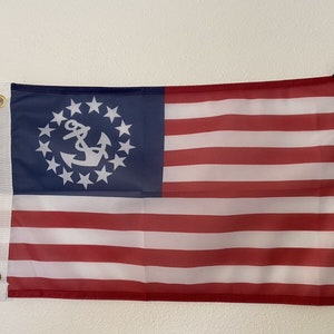 May include: A red, white, and blue American flag with a white anchor and 13 stars in a circle on the blue field. The flag is hanging on a white wall.