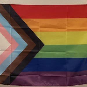 May include: A flag with a rainbow design and a white, pink, light blue, brown, and black chevron. The flag has a red, orange, yellow, green, blue, and purple striped background. The flag is hanging on a wall.