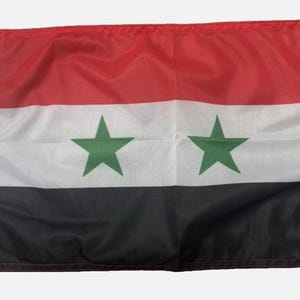 May include: Syrian flag with red, white, and black horizontal stripes. Two green five-pointed stars are centered on the white stripe. The flag has a white reinforced edge with brass grommets.