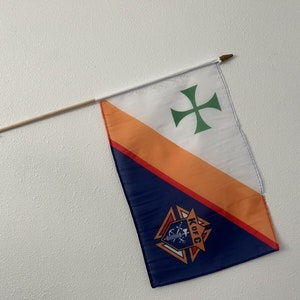 May include: A small, blue, orange, and white flag with a green cross and a Knights of Columbus logo. The flag is attached to a wooden stick.