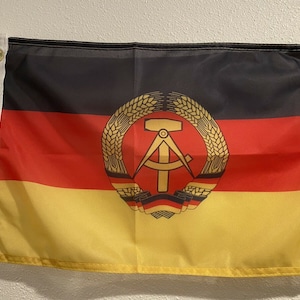 May include: A black, red, and yellow flag with a gold emblem featuring a compass, hammer, and wheat stalks. The emblem is centered on the flag.