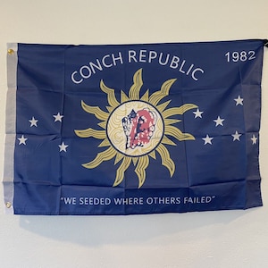 May include: A blue flag with a yellow sun and a conch shell in the center. The text "CONCH REPUBLIC 1982" is printed in white at the top of the flag. The text "WE SEEDED WHERE OTHERS FAILED" is printed in white at the bottom of the flag.