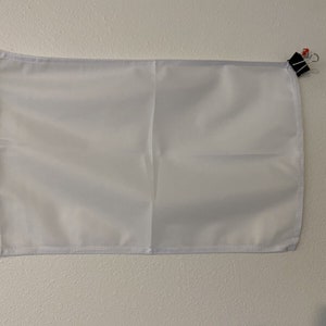 May include: A plain white banner with gold grommets at the top and bottom. The banner is hanging on a white wall with two black binder clips holding the top corner.