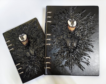 Könnte beinhalten: Zwei schwarze Notizbücher mit einem 3D-Venom-Charakterdesign. Die Notizbücher haben einen schwarzen Einband mit einem strukturierten Design und eine silberne Spiralbindung.