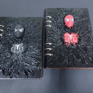 May include: Two black spiral notebooks with a textured, black, web-like design. One notebook features a black and white Spider-Man figure, while the other features a red and black Spider-Man figure.