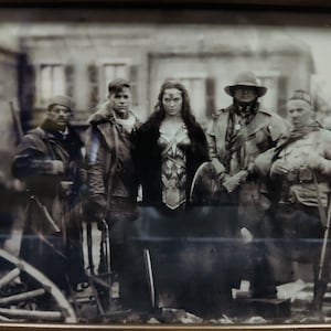 Peut inclure: Une photo en noir et blanc d'un groupe de personnes debout devant un bâtiment. Les personnes sont habillées en uniformes militaires et tiennent des armes. Une femme se tient au centre du groupe et porte un costume avec un plastron et une jupe.