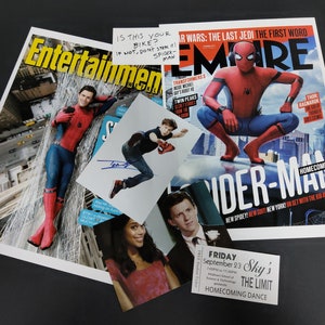 May include: A red and blue Spider-Man costume with a red mask. The costume is on a magazine cover with the title "Spider-Man Homecoming". The magazine is on a black surface with a signed photo of Spider-Man and a ticket for a homecoming dance.